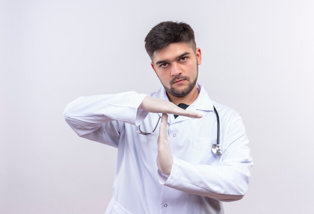 Joven apuesto médico con bata médica blanca, guantes médicos blancos y un estetoscopio que muestra el tiempo de descanso con las manos sobre la pared blanca