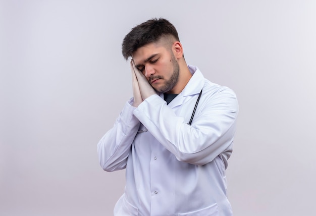 Joven apuesto médico con bata médica blanca, guantes médicos blancos y estetoscopio quiere dormir de pie sobre una pared blanca