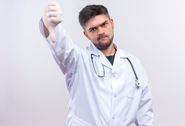 Joven apuesto médico con bata médica blanca, guantes médicos blancos y un estetoscopio con reproche haciendo malos pulgares abajo de pie sobre la pared blanca