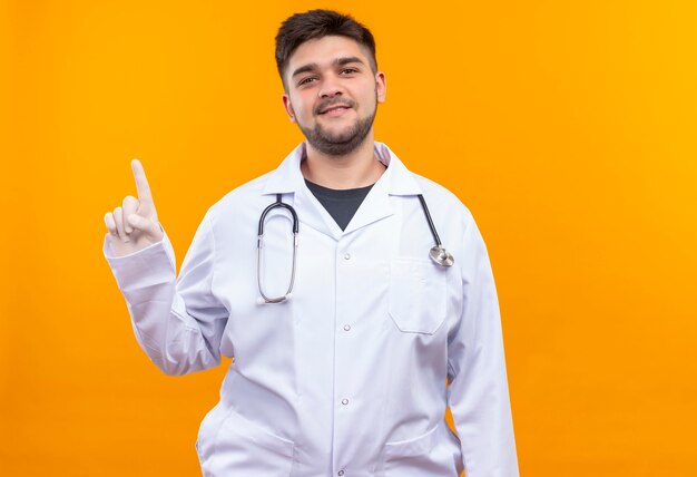 Joven apuesto médico con bata médica blanca, guantes médicos blancos y un estetoscopio sonriendo apuntando hacia arriba con el dedo índice de pie sobre la pared naranja