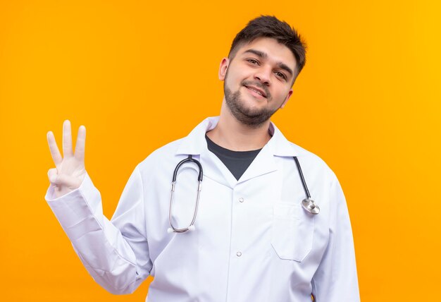 Joven apuesto médico con bata médica blanca guantes médicos blancos y un estetoscopio sonriendo mostrando tres con los dedos de pie sobre la pared naranja