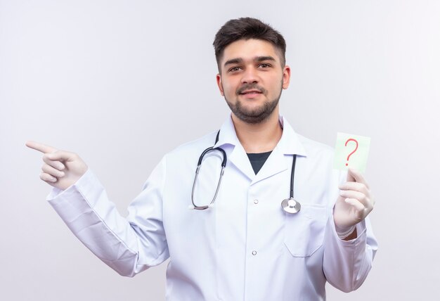 Joven apuesto médico con bata médica blanca, guantes médicos blancos y un estetoscopio sonriendo sosteniendo un cartel de pregunta y apuntando a la derecha sobre la pared blanca