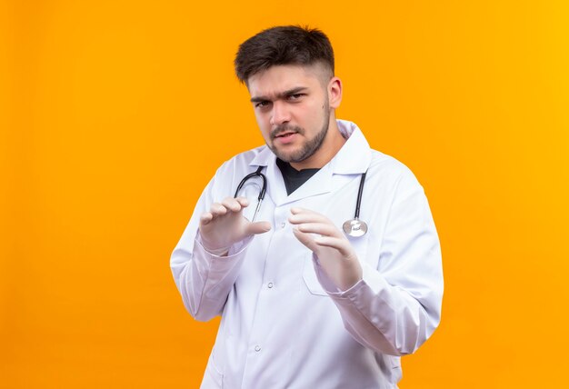 Joven apuesto médico con bata médica blanca, guantes médicos blancos y estetoscopio por temor a algo parado sobre la pared naranja