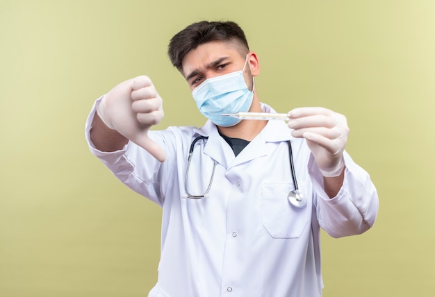 Joven apuesto médico con máscara médica azul bata médica blanca guantes médicos blancos y un estetoscopio apuntando al termómetro tristemente por la temperatura haciendo infelices pulgares hacia abajo de pie