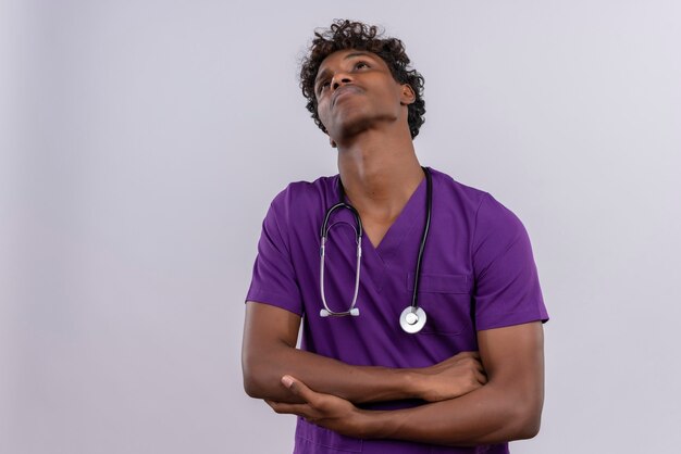 Un joven apuesto médico de piel oscura con cabello rizado vistiendo uniforme violeta con estetoscopio mirando hacia arriba sosteniendo el codo