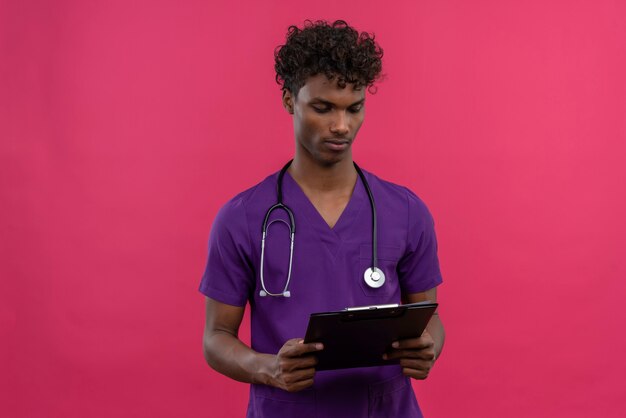 Un joven apuesto médico de piel oscura con cabello rizado vistiendo uniforme violeta con estetoscopio mirando el portapapeles