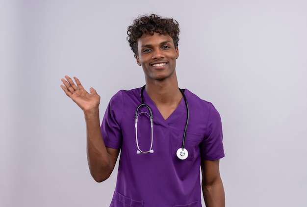 Un joven apuesto médico de piel oscura con cabello rizado vistiendo uniforme violeta con estetoscopio mostrando adiós