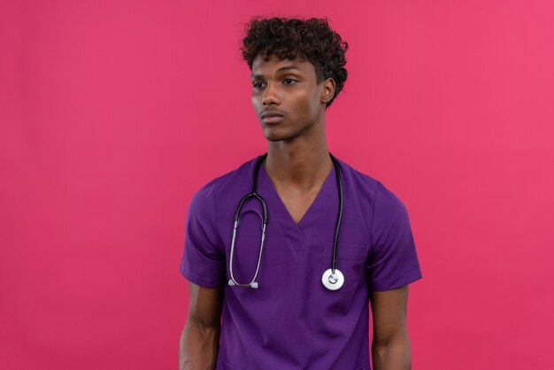 Un joven apuesto médico de piel oscura con cabello rizado vistiendo uniforme violeta con estetoscopio de pie y con aspecto cansado