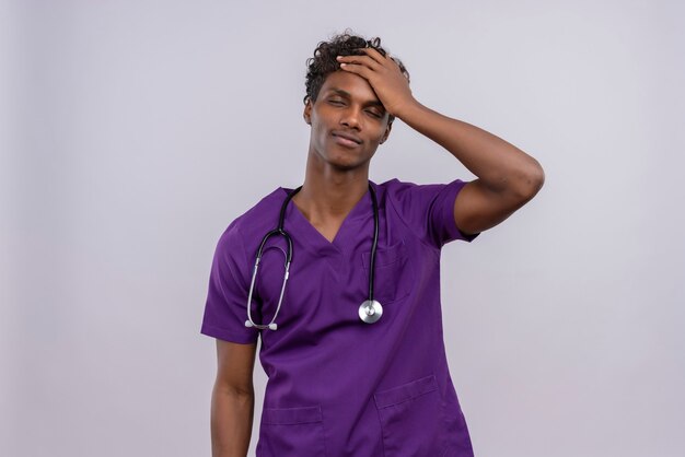 Un joven apuesto médico de piel oscura con cabello rizado vistiendo uniforme violeta con estetoscopio sintiéndose mal teniendo presión física de alta temperatura manteniendo los ojos cerrados tocando la frente con pa