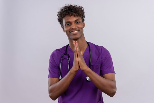 Un joven apuesto médico de piel oscura con cabello rizado vistiendo uniforme violeta con estetoscopio tomados de la mano juntos en el gesto de agradecimiento