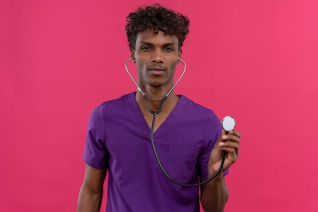 Un joven apuesto médico de piel oscura con cabello rizado vistiendo uniforme violeta mostrando estetoscopio mientras