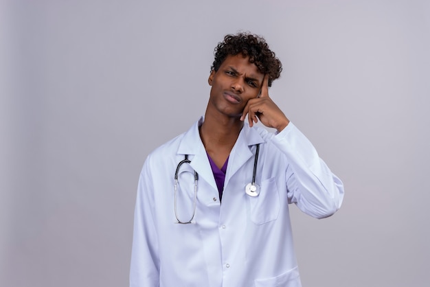Un joven apuesto médico de piel oscura con pelo rizado vestido con bata blanca con estetoscopio manteniendo la mano en la cabeza mientras piensa