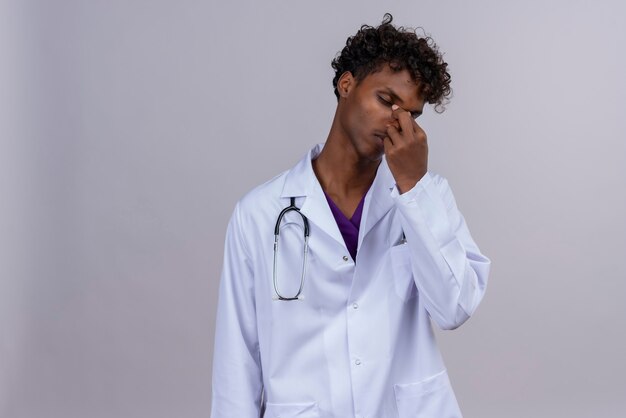 Un joven apuesto médico de piel oscura con pelo rizado vistiendo bata blanca con estetoscopio sosteniendo la nariz con los dedos