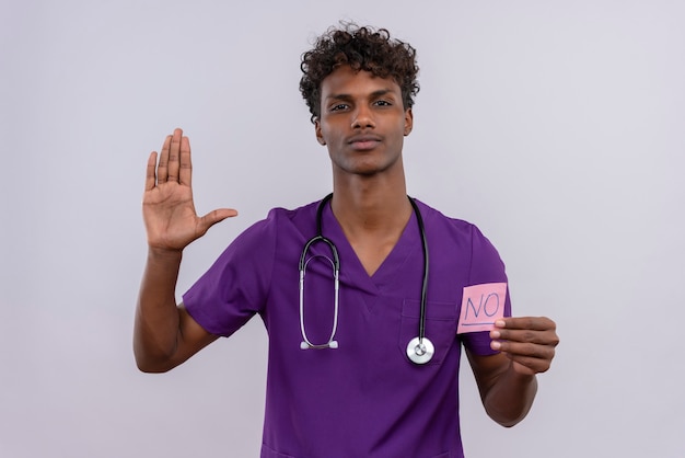 Un joven apuesto médico varón de piel oscura con cabello rizado vistiendo uniforme violeta con estetoscopio mostrando una tarjeta de papel con la palabra no