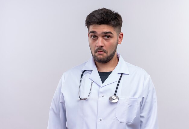 Joven apuesto médico vistiendo una bata médica blanca y un estetoscopio mirando sorprendido de pie sobre la pared blanca