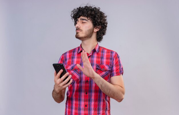 Un joven apuesto con el pelo rizado en camisa a cuadros cerrando los ojos sosteniendo el teléfono móvil