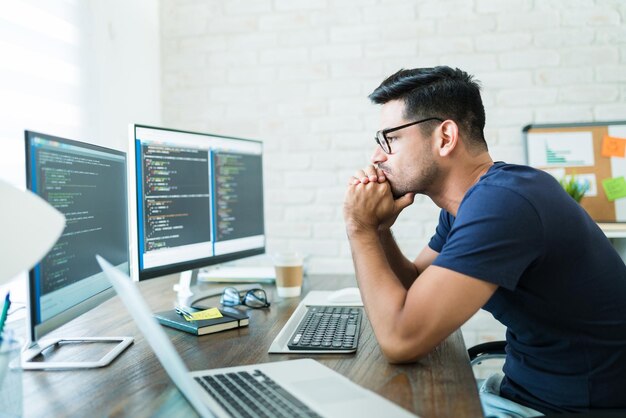 Un joven y apuesto programador pensativo que analiza códigos de software en pantallas de computadora mientras trabaja desde casa