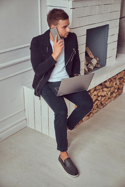 Un joven apuesto y relajado con ropa elegante y reloj de pulsera sentado en una chimenea. trabajando en su computadora portátil y hablando por teléfono.