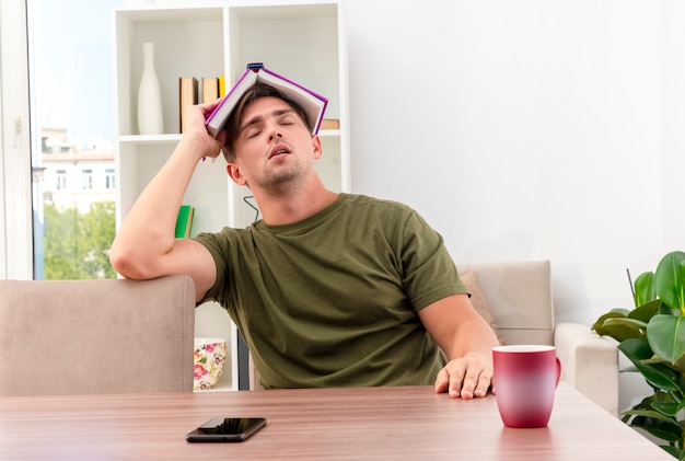 Joven apuesto rubio soñoliento se sienta a la mesa con una taza y un teléfono sosteniendo un libro sobre la cabeza con los ojos cerrados dentro de la sala de estar