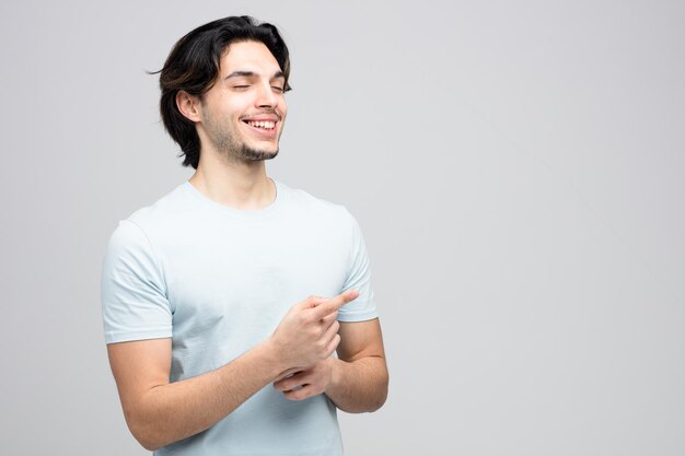 un joven apuesto sonriente apuntando a un lado con los ojos cerrados aislado en un fondo blanco con espacio para copiar