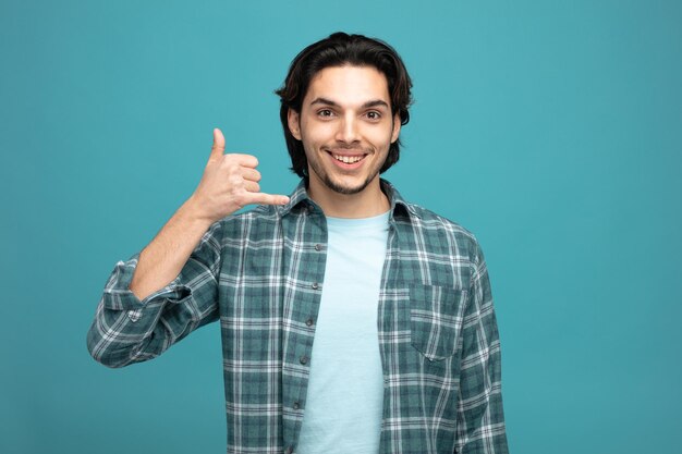 un joven apuesto sonriente mirando a la cámara mostrando el gesto de llamarme aislado de fondo azul