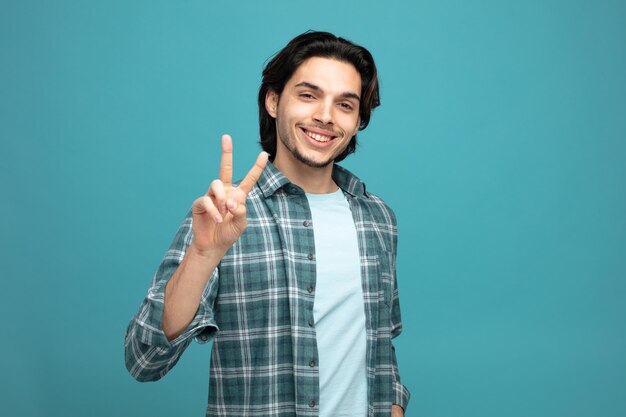 un joven apuesto sonriente mirando una cámara que muestra el signo de la paz aislado de fondo azul