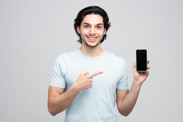 un joven apuesto sonriente mirando una cámara que muestra un teléfono móvil apuntándolo aislado de fondo blanco