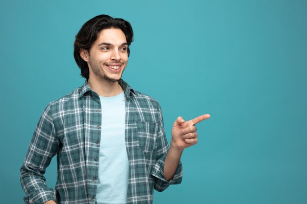 un joven apuesto sonriente mirando y señalando a un lado aislado en un fondo azul con espacio para copiar