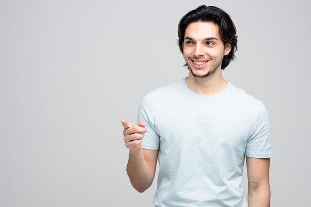 un joven apuesto sonriente mirando y señalando a un lado aislado de fondo blanco con espacio para copiar