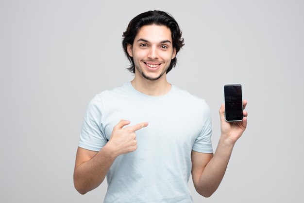 un joven apuesto sonriente que muestra un teléfono móvil apuntándolo mirando a la cámara aislada de fondo blanco