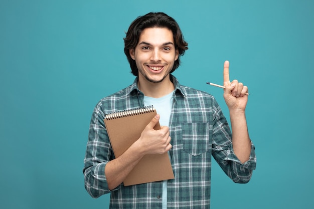 un joven apuesto sonriente sosteniendo un bloc de notas y un lápiz mirando a la cámara apuntando hacia arriba aislado de fondo azul
