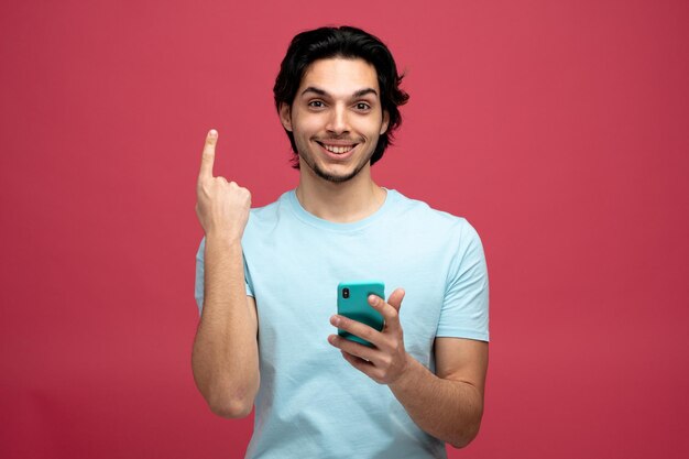 un joven apuesto sonriente sosteniendo un teléfono móvil mirando a la cámara apuntando hacia arriba aislado de fondo rojo