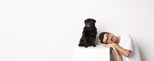 Foto gratuita un joven apuesto yacía la cabeza cerca de un lindo perro negro sonriendo y mirando el fondo blanco de la cámara