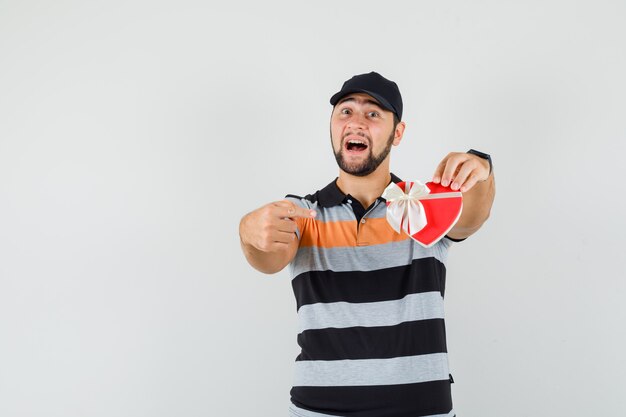 Joven apuntando al cuadro actual en camiseta, gorra y mirando alegre. vista frontal.