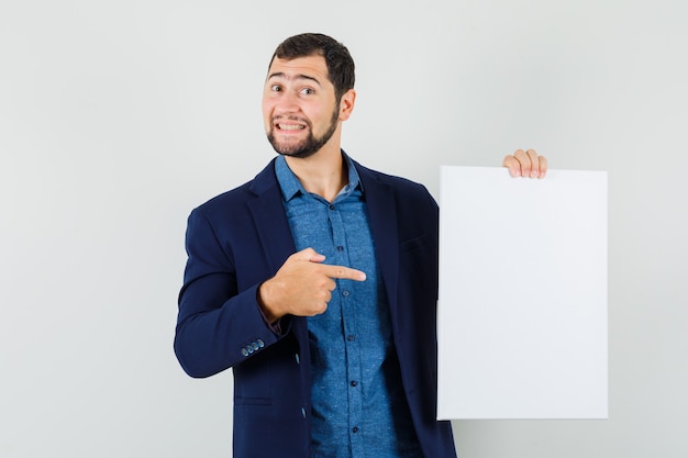 Foto gratuita joven apuntando al lienzo en blanco en camisa, chaqueta y mirando alegre, vista frontal.