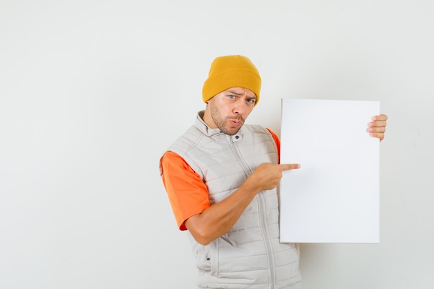 Joven apuntando al lienzo en blanco en camiseta, chaqueta, sombrero, vista frontal.