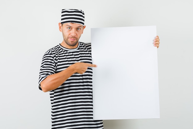 Joven apuntando al lienzo en blanco con sombrero de camiseta a rayas y mirando positivo