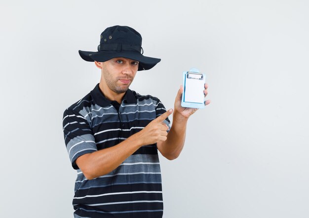 Joven apuntando al mini portapapeles en camiseta, sombrero y mirando positivo, vista frontal.