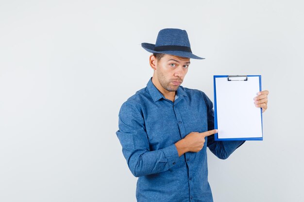 Joven apuntando al portapapeles con camisa azul, sombrero y mirando serio, vista frontal.