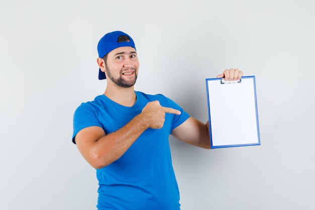 Joven apuntando al portapapeles en camiseta azul y gorra y mirando alegre