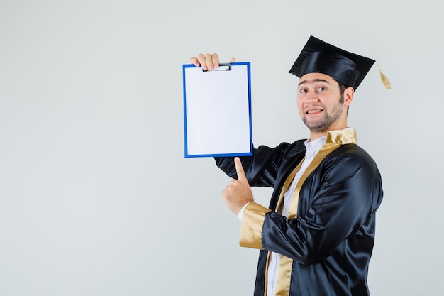 Foto gratuita joven apuntando al portapapeles en uniforme de posgrado y mirando confiado, vista frontal.