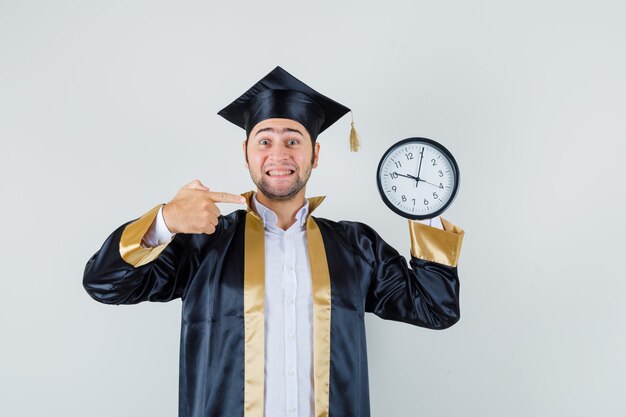 Joven apuntando al reloj de pared en uniforme de posgrado y mirando alegre, vista frontal.