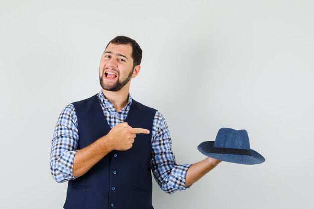 Foto gratuita joven apuntando al sombrero con camisa, chaleco y mirando confiado, vista frontal.