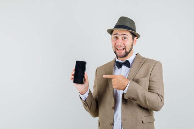 Joven apuntando al teléfono móvil en traje, sombrero y mirando alegre, vista frontal.