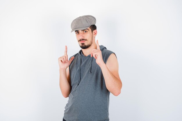 Joven apuntando hacia arriba en camiseta gris y gorra y mirando serio