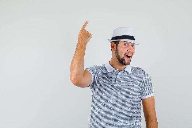 Joven apuntando hacia arriba con camiseta a rayas, sombrero y aspecto alegre. vista frontal.