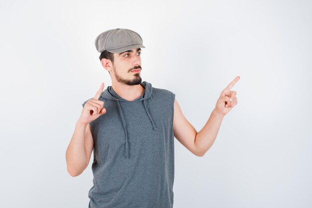 Joven apuntando hacia arriba y hacia la derecha con los dedos índices en camiseta gris y gorra y mirando serio