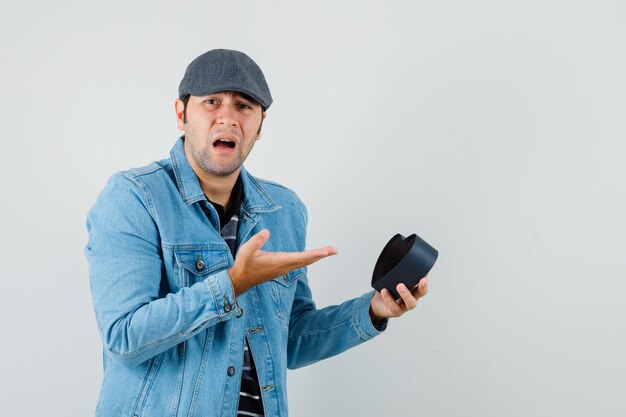 Joven apuntando a caja de regalo en chaqueta, gorra y mirando insatisfecho.