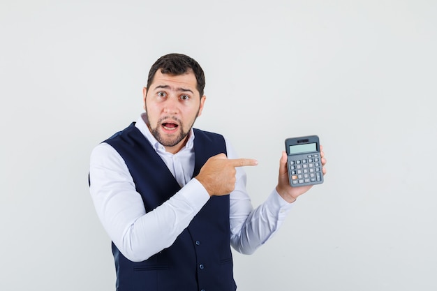 Joven apuntando a la calculadora en camisa, chaleco y mirando desconcertado