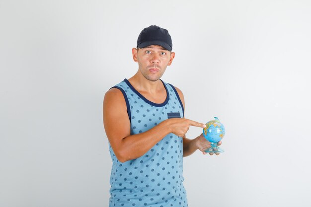 Joven apuntando con el dedo al globo en camiseta azul con gorra y mirando confundido